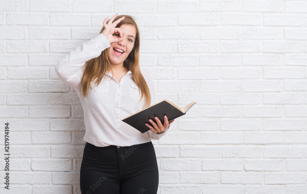 Young adult woman standing over white brick wall reading a book with happy face smiling doing ok sign with hand on eye looking through fingers
