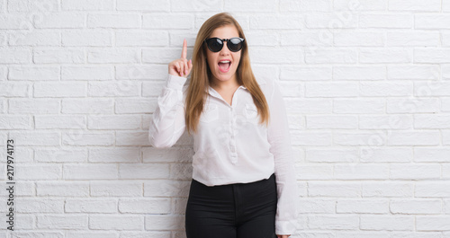 Young adult business woman standing over white brick wall wearing sunglasses surprised with an idea or question pointing finger with happy face, number one © Krakenimages.com