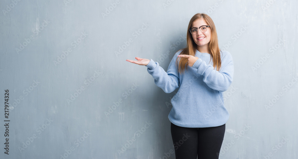 Young adult woman over grey grunge wall wearing glasses amazed and smiling to the camera while presenting with hand and pointing with finger.