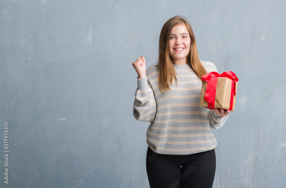 Young adult woman over grey grunge wall holding a present screaming proud and celebrating victory and success very excited, cheering emotion Young adult woman over grey grunge wall holding a present screaming proud and celebrating victory and success very excited, cheering emotion