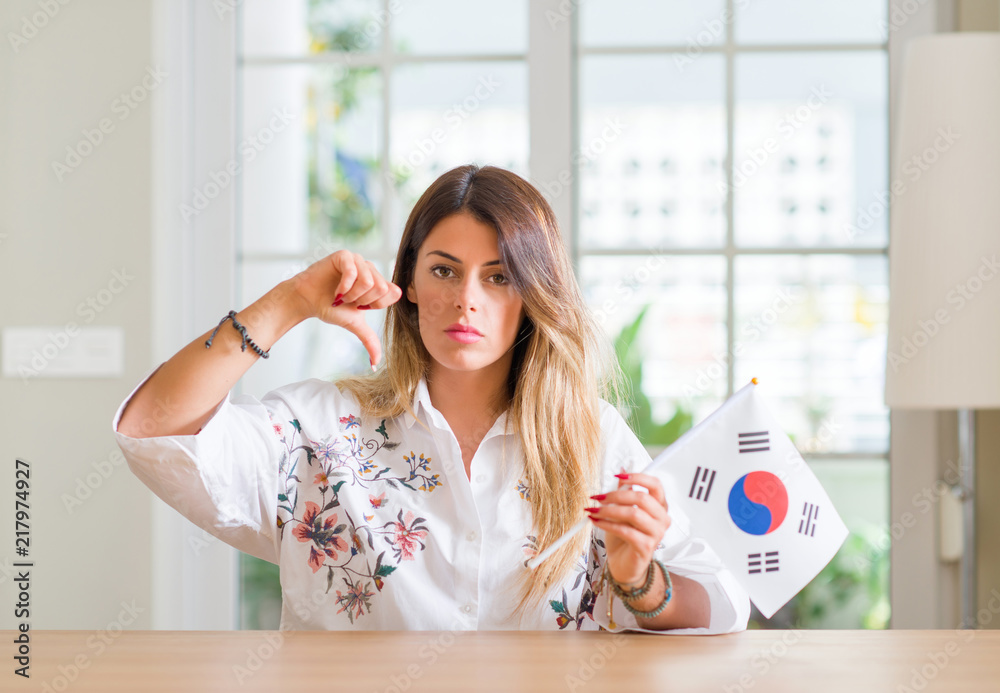Young woman at home holding flag of South Korea with angry face, negative sign showing dislike with thumbs down, rejection concept Young woman at home holding flag of South Korea with angry face, negative sign showing dislike with thumbs down, rejection concept