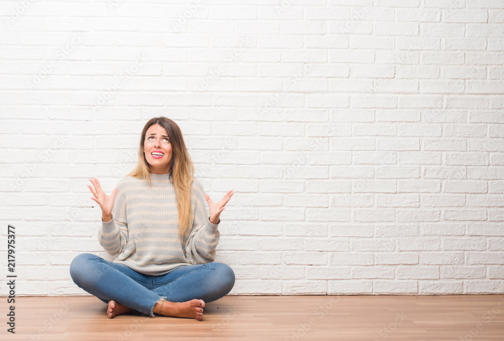 Young adult woman sitting on the floor over white brick wall at home crazy and mad shouting and yelling with aggressive expression and arms raised. Frustration concept.