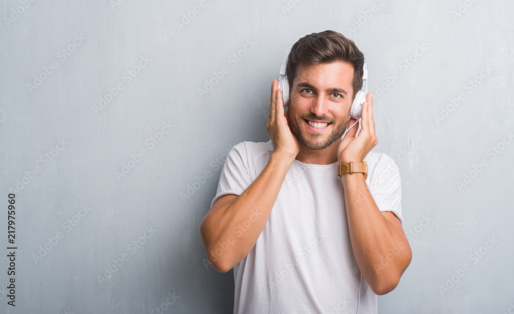 Handsome young man over grey grunge wall wearing headphones listening to music with a happy face standing and smiling with a confident smile showing teeth Handsome young man over grey grunge wall wearing headphones listening to music with a happy face standing and smiling with a confident smile showing teeth