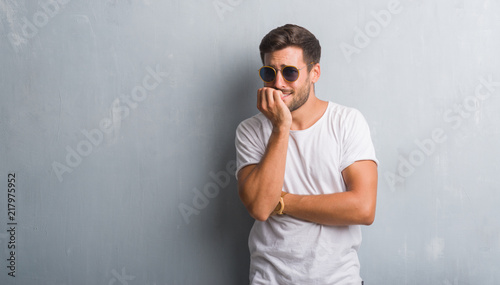 Handsome young man over grey grunge wall wearing sunglasses looking stressed and nervous with hands on mouth biting nails. Anxiety problem.