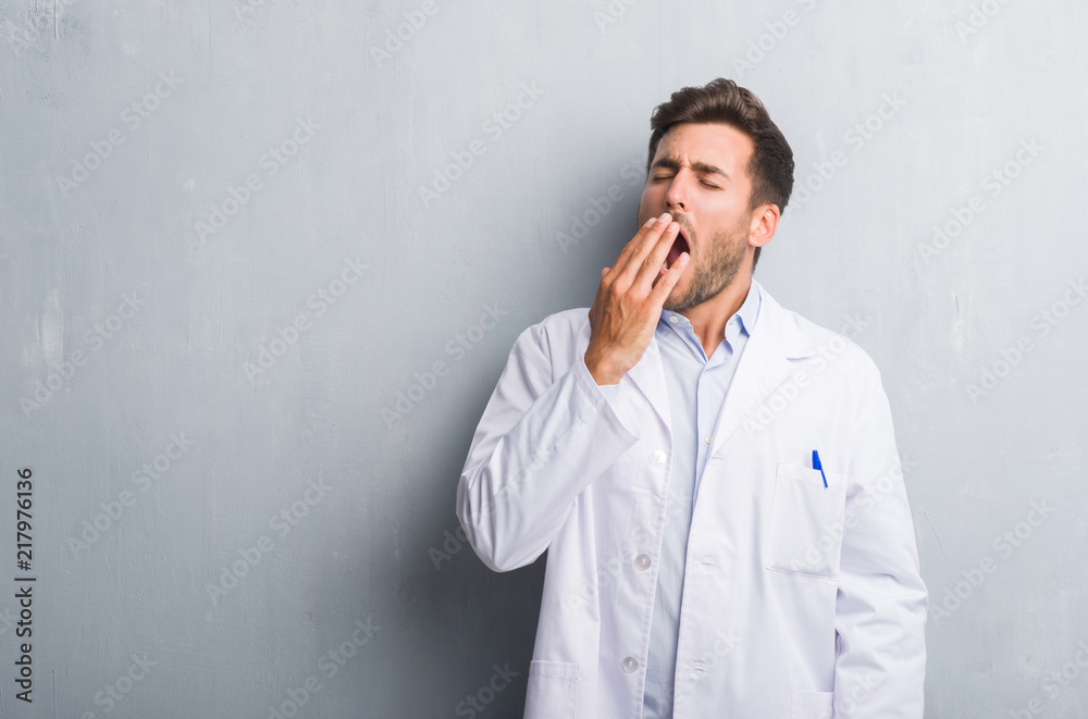 Handsome young professional man over grey grunge wall wearing white coat bored yawning tired covering mouth with hand. Restless and sleepiness. Handsome young professional man over grey grunge wall wearing white coat bored yawning tired covering mouth with hand. Restless and sleepiness.