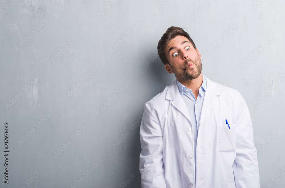 Handsome young professional man over grey grunge wall wearing white coat making fish face with lips, crazy and comical gesture. Funny expression. Handsome young professional man over grey grunge wall wearing white coat making fish face with lips, crazy and comical gesture. Funny expression.