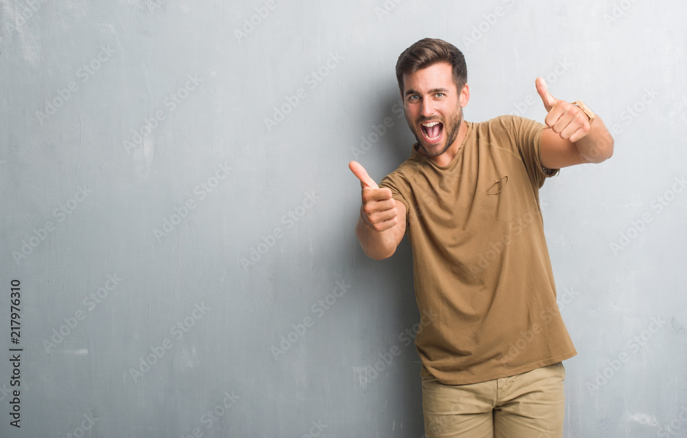 Handsome young man over grey grunge wall approving doing positive gesture with hand, thumbs up smiling and happy for success. Looking at the camera, winner gesture. Handsome young man over grey grunge wall approving doing positive gesture with hand, thumbs up smiling and happy for success. Looking at the camera, winner gesture.