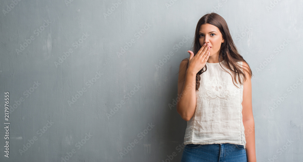 Fototapeta premium Young brunette woman over grunge grey wall cover mouth with hand shocked with shame for mistake, expression of fear, scared in silence, secret concept