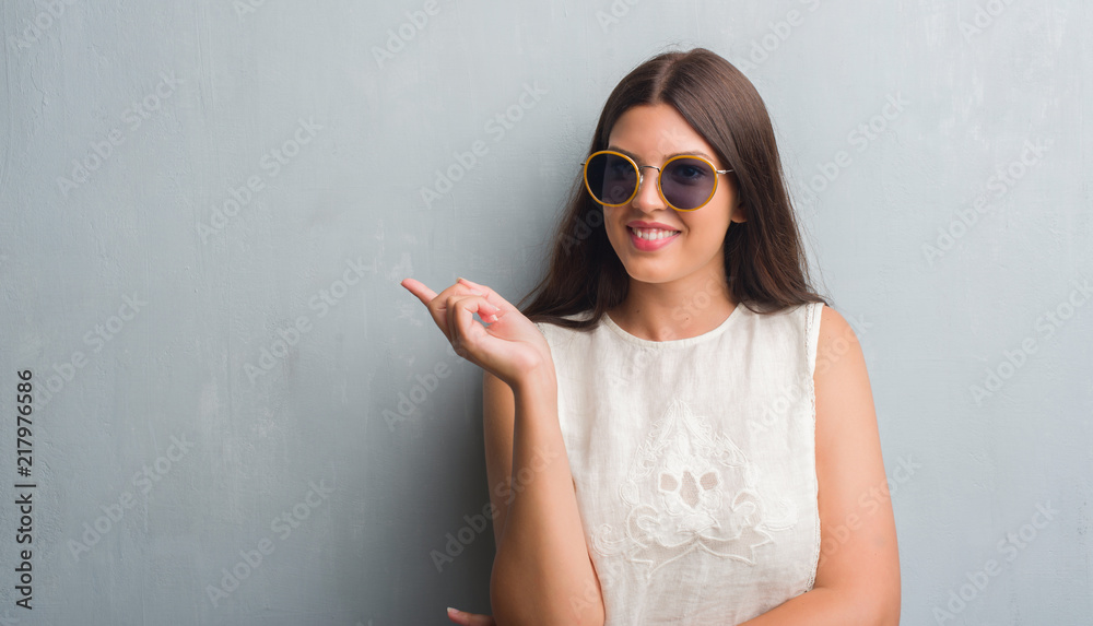 Young brunette woman over grunge grey wall wearing retro sunglasses very happy pointing with hand and finger to the side Young brunette woman over grunge grey wall wearing retro sunglasses very happy pointing with hand and finger to the side