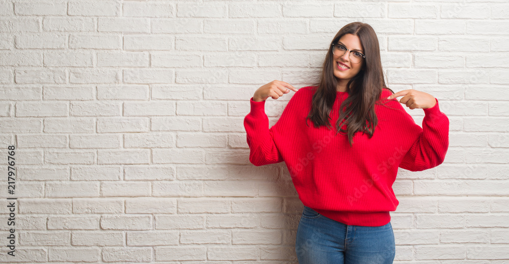 Young brunette woman standing over white brick wall looking confident with smile on face, pointing oneself with fingers proud and happy.