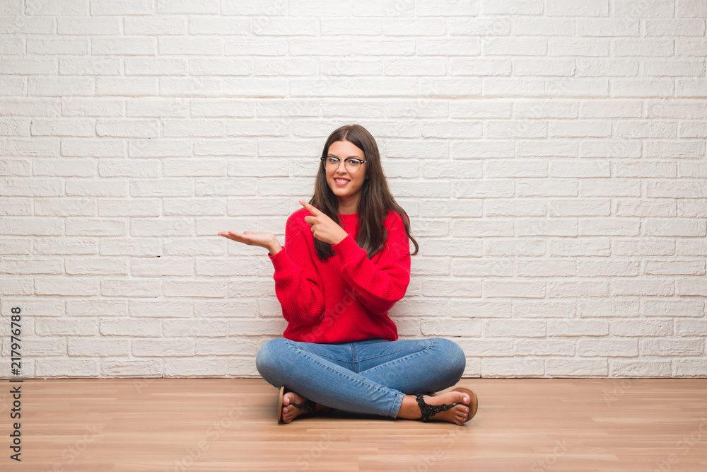 Young brunette woman sitting on the floor over white brick wall very happy pointing with hand and finger Young brunette woman sitting on the floor over white brick wall very happy pointing with hand and finger
