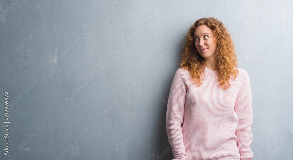 Young redhead woman over grey grunge wall wearing pink sweater smiling looking side and staring away thinking. Young redhead woman over grey grunge wall wearing pink sweater smiling looking side and staring away thinking.