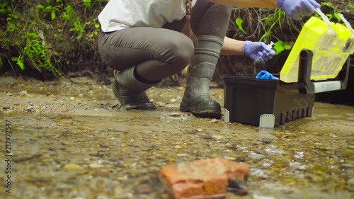 Woman scientist environmentalist sitting near the creek. She opening a tool box, taking sample of water and pouring it into the test tube.