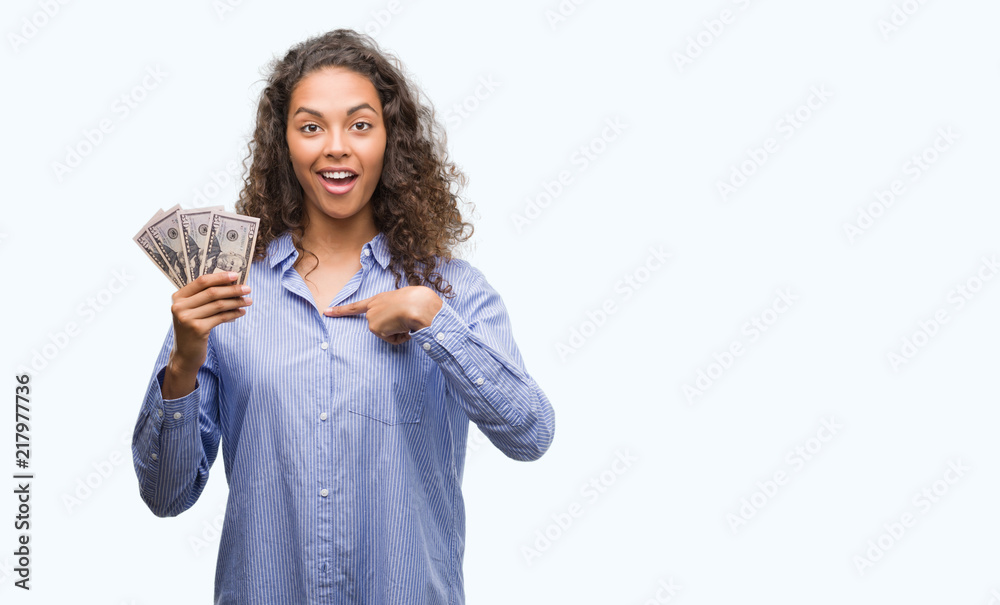 Young hispanic woman holding dollars with surprise face pointing finger to himself Young hispanic woman holding dollars with surprise face pointing finger to himself