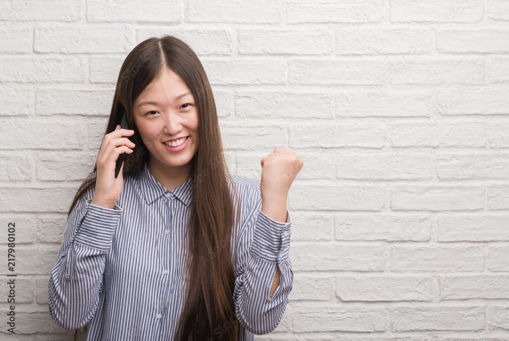 Young Chinese woman over brick wall talking on the phone screaming proud and celebrating victory and success very excited, cheering emotion Young Chinese woman over brick wall talking on the phone screaming proud and celebrating victory and success very excited, cheering emotion