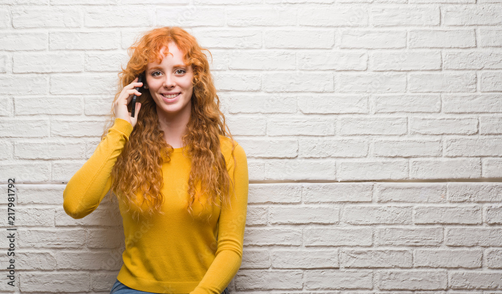 Young redhead woman standing over brick wall talking on the phone with a happy face standing and smiling with a confident smile showing teeth Young redhead woman standing over brick wall talking on the phone with a happy face standing and smiling with a confident smile showing teeth