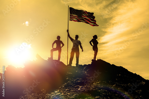 silhouettes of three happy man on a mountain top, with flag america, on sunset background. success and achievements. teamwork.
