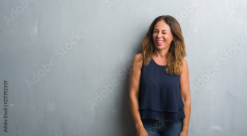 Middle age hispanic woman standing over grey grunge wall winking looking at the camera with sexy expression, cheerful and happy face. © Krakenimages.com