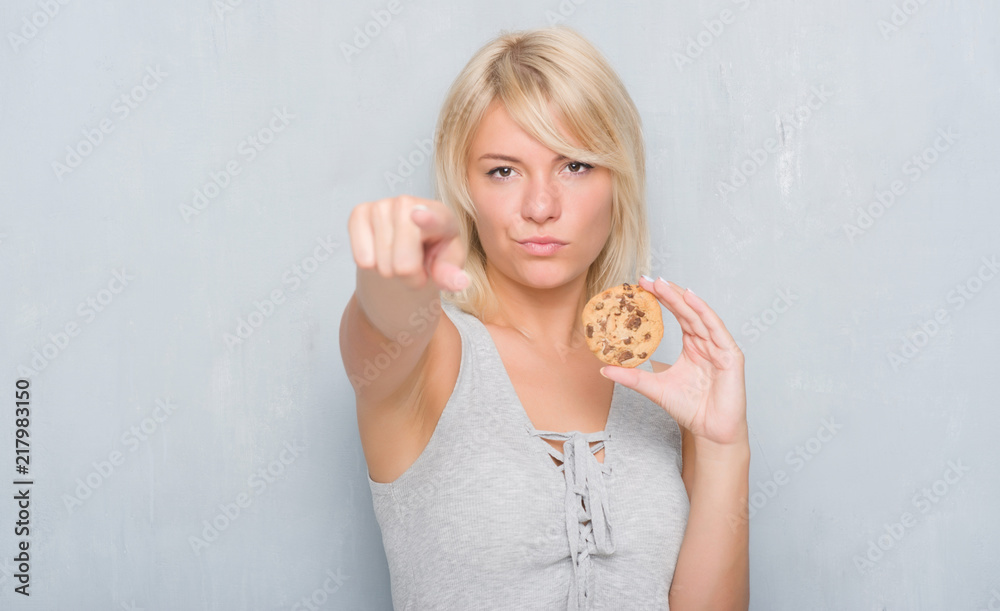 Caucasian adult woman over grey grunge wall eating chocolate cooky pointing with finger to the camera and to you, hand sign, positive and confident gesture from the front Caucasian adult woman over grey grunge wall eating chocolate cooky pointing with finger to the camera and to you, hand sign, positive and confident gesture from the front