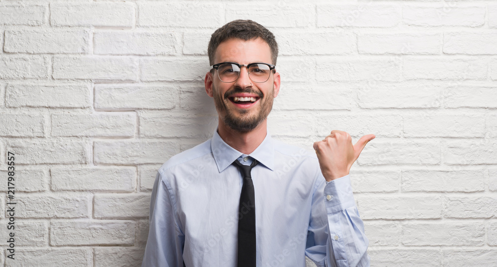 Young adult business man standing over white brick wall pointing and showing with thumb up to the side with happy face smiling Young adult business man standing over white brick wall pointing and showing with thumb up to the side with happy face smiling