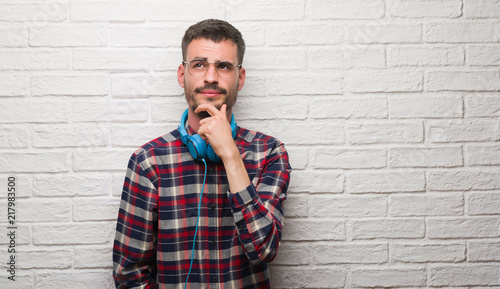 Young adult man over brick wall wearing headphones serious face thinking about question, very confused idea