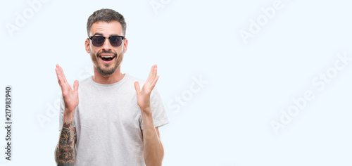 Young tattooed adult man wearing sunglasses very happy and excited, winner expression celebrating victory screaming with big smile and raised hands