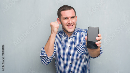 Young caucasian man over grey grunge wall showing blank screen of smartphone annoyed and frustrated shouting with anger, crazy and yelling with raised hand, anger concept © Krakenimages.com