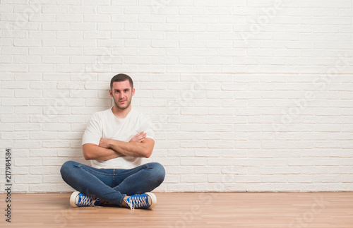 Young caucasian man sitting on the floor over white brick wall skeptic and nervous, disapproving expression on face with crossed arms. Negative person. © Krakenimages.com