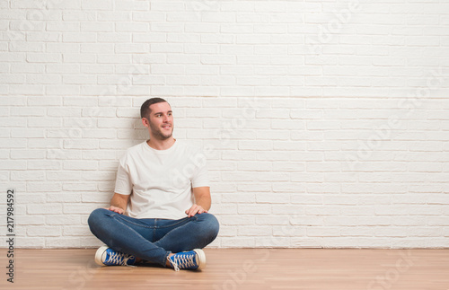 Young caucasian man sitting on the floor over white brick wall looking away to side with smile on face, natural expression. Laughing confident. © Krakenimages.com