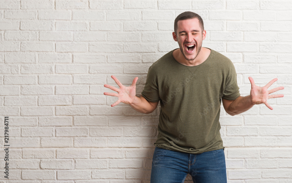 Young caucasian man standing over white brick wall celebrating mad and crazy for success with arms raised and closed eyes screaming excited. Winner concept Young caucasian man standing over white brick wall celebrating mad and crazy for success with arms raised and closed eyes screaming excited. Winner concept