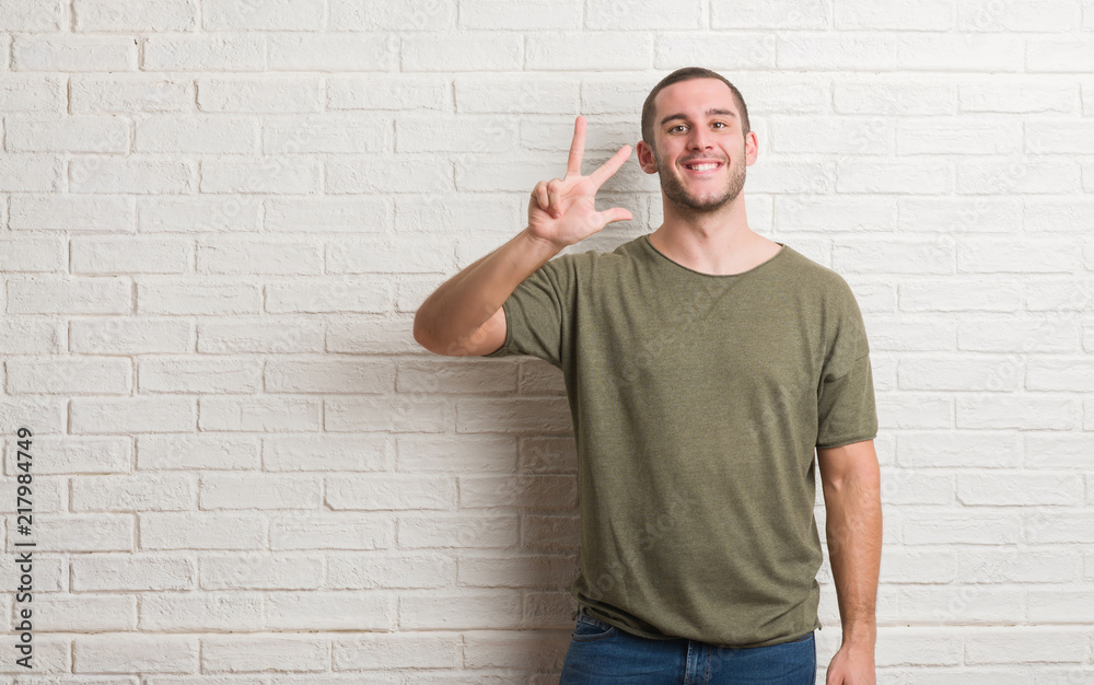 Young caucasian man standing over white brick wall showing and pointing up with fingers number three while smiling confident and happy. Young caucasian man standing over white brick wall showing and pointing up with fingers number three while smiling confident and happy.