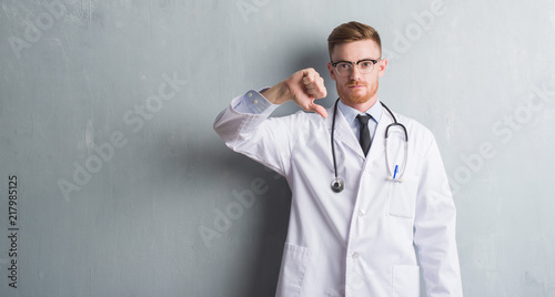 Young redhead doctor man over grey grunge wall with angry face, negative sign showing dislike with thumbs down, rejection concept