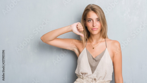 Beautiful young woman standing over grunge grey wall with angry face, negative sign showing dislike with thumbs down, rejection concept