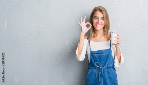 Beautiful young woman over grunge grey wall driking a glass of milk doing ok sign with fingers, excellent symbol