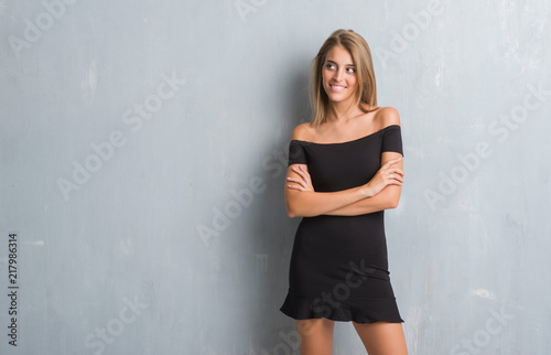 Beautiful young woman standing over grunge grey wall wearing elegant dress smiling looking side and staring away thinking.