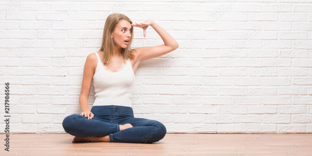 Beautiful young woman sitting on the floor at home very happy and smiling looking far away with hand over head. Searching concept. Beautiful young woman sitting on the floor at home very happy and smiling looking far away with hand over head. Searching concept.