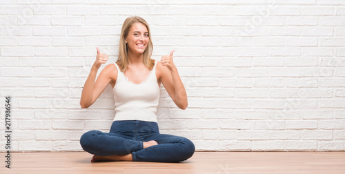Beautiful young woman sitting on the floor at home success sign doing positive gesture with hand, thumbs up smiling and happy. Looking at the camera with cheerful expression, winner gesture.