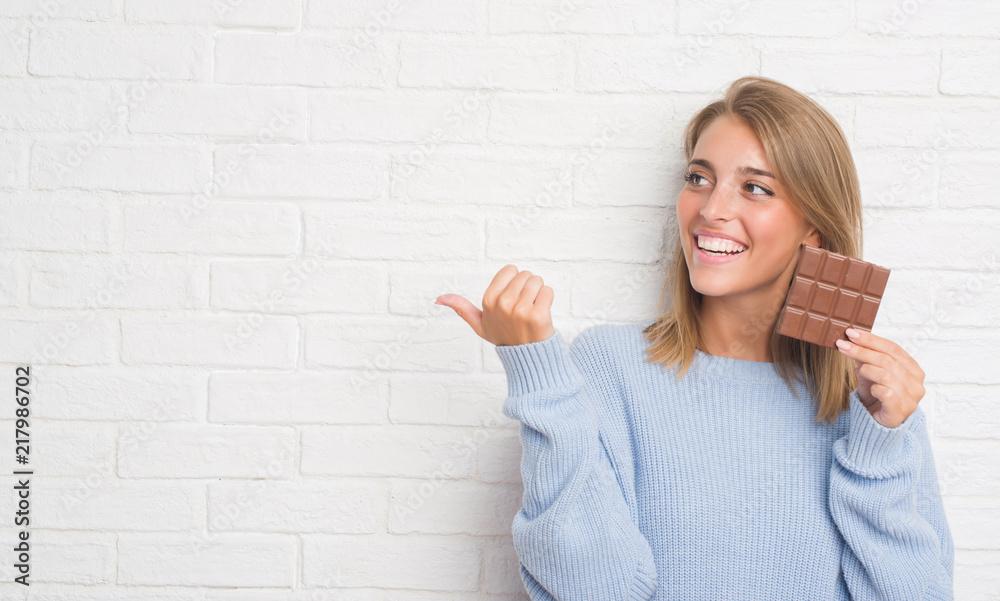 Beautiful young woman over white brick wall eating chocolate bar pointing and showing with thumb up to the side with happy face smiling Beautiful young woman over white brick wall eating chocolate bar pointing and showing with thumb up to the side with happy face smiling