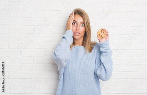 Beautiful young woman over white brick wall eating chocolate chip cooky stressed with hand on head, shocked with shame and surprise face, angry and frustrated. Fear and upset for mistake. © Krakenimages.com