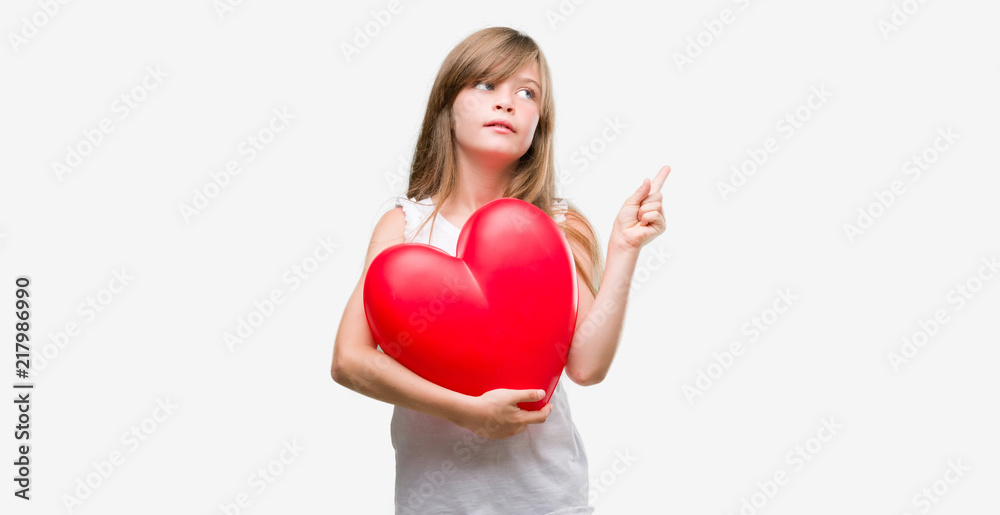 Young blonde toddler holding a red heart very happy pointing with hand and finger to the side Young blonde toddler holding a red heart very happy pointing with hand and finger to the side