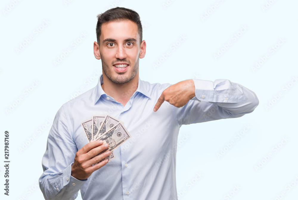 Handsome young man holding money with surprise face pointing finger to himself