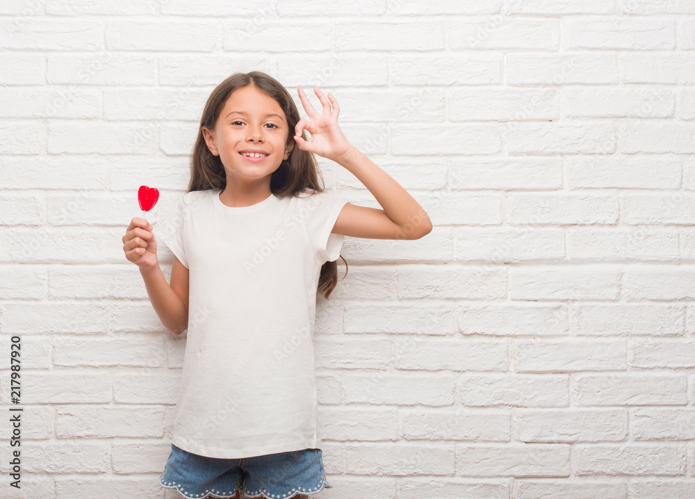 Young hispanic kid over white brick wall eating red heart lollipop candy doing ok sign with fingers, excellent symbol Young hispanic kid over white brick wall eating red heart lollipop candy doing ok sign with fingers, excellent symbol