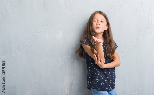 Young hispanic kid over grunge grey wall looking at the camera blowing a kiss with hand on air being lovely and sexy. Love expression.