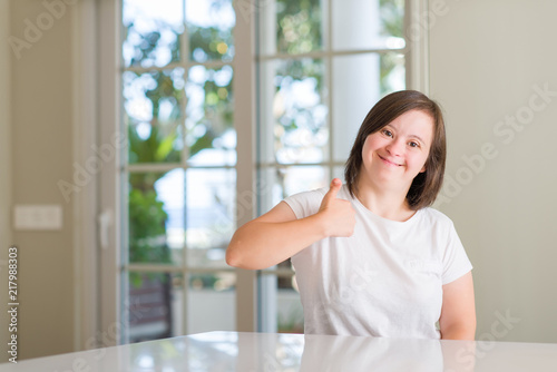 Down syndrome woman at home doing happy thumbs up gesture with hand. Approving expression looking at the camera with showing success.