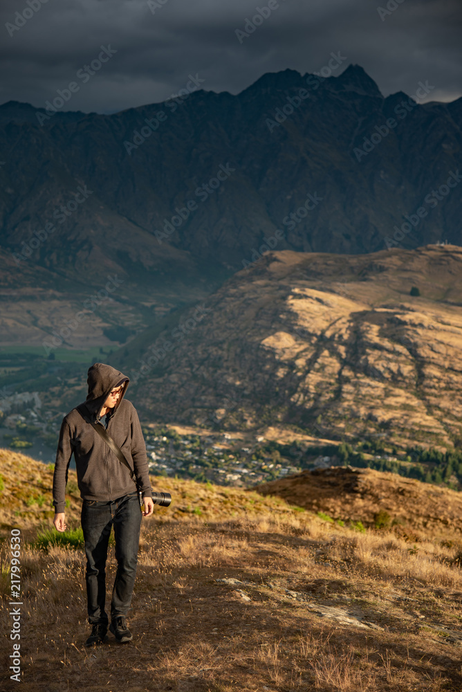 Young male photographer walking on Queenstown hill with mountain scenery in the background during golden hour sunset in Queenstown, South Island, New Zealand. Travel and photography concepts