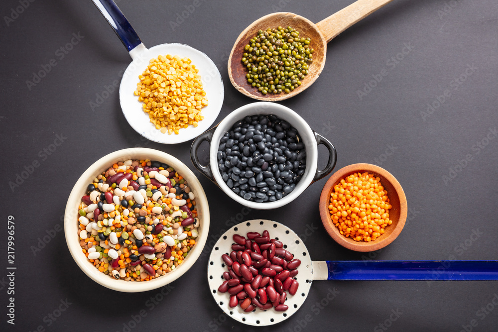 Top view of fflat lay of assortment of legumes pulses on black tabletop background, in scoop, enameled bowl and ladles.