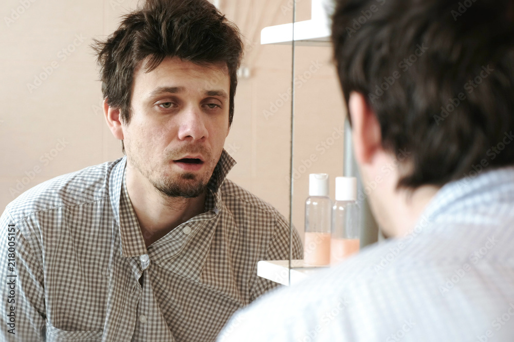 Fototapeta premium Tired man who has just woken up looks at his reflection in the mirror and sees his scruffy appearance