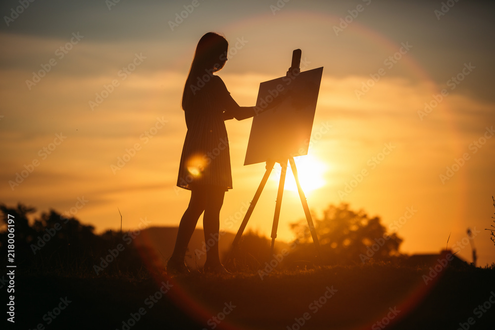 Obraz premium Silhouette of a blonde girl artist. Lady paints a painting on the canvas with the help of paints. A wooden easel keeps the picture. Summer is a sunny day, sunset