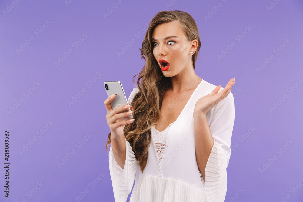 Photo of shocked european woman 20s with long curly hairstyle wearing dress holding and looking at mobile phone, isolated over violet background Photo of shocked european woman 20s with long curly hairstyle wearing dress holding and looking at mobile phone, isolated over violet background