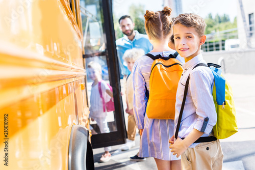 adorable schoolboy entering school bus with classmates while teacher standing near door
